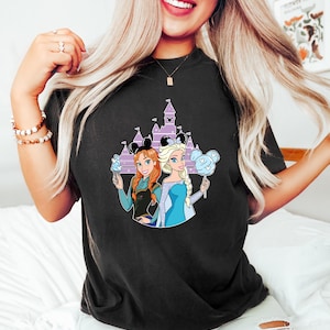 May include: Black t-shirt featuring a graphic of two cartoon princesses with ice cream and cotton candy. The princesses are in front of a purple castle with Mickey Mouse ears. The shirt is a comfortable, casual style.
