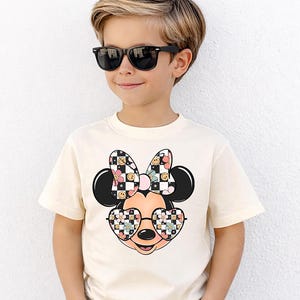 Puede incluir: Una camiseta de manga corta de color crema con un gr&aacute;fico de un personaje de rat&oacute;n de dibujos animados que lleva gafas en forma de coraz&oacute;n. El lazo y las gafas del personaje est&aacute;n decorados con un patr&oacute;n de cuadros blanco y negro, flores rosas y caras sonrientes amarillas.