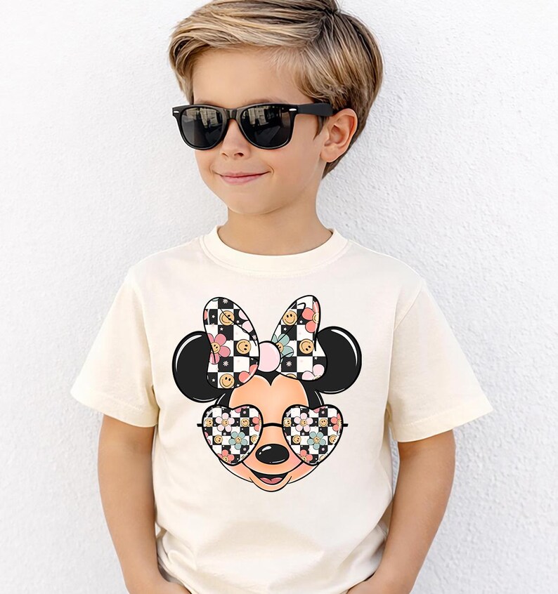 Puede incluir: Una camiseta de manga corta de color crema con un gr&aacute;fico de un personaje de rat&oacute;n de dibujos animados que lleva gafas en forma de coraz&oacute;n. El lazo y las gafas del personaje est&aacute;n decorados con un patr&oacute;n de cuadros blanco y negro, flores rosas y caras sonrientes amarillas.