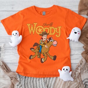 May include: Orange t-shirt featuring Sheriff Woody riding a horse, with the text "Sheriff Woody" in a Western-style font. The shirt is surrounded by three white ghost plushies, perfect for Halloween or a Toy Story fan.