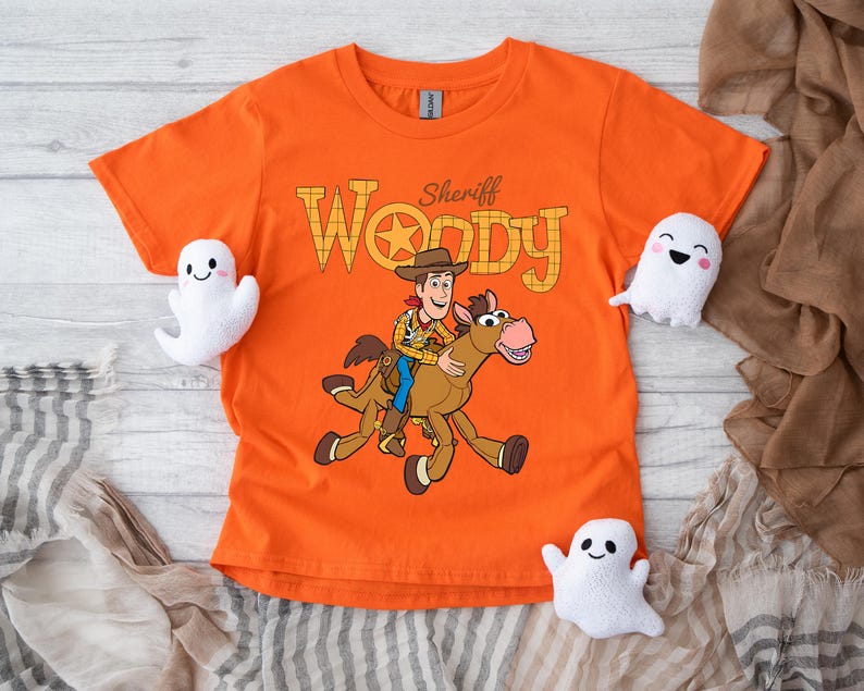 May include: Orange t-shirt featuring Sheriff Woody riding a horse, with the text "Sheriff Woody" in a Western-style font. The shirt is surrounded by three white ghost plushies, perfect for Halloween or a Toy Story fan.