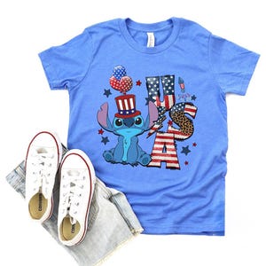 K&ouml;nnte beinhalten: Hellblaues T-Shirt mit einer Cartoonfigur, die einen patriotischen Hut tr&auml;gt und das Wort "USA" in Rot, Wei&szlig; und Blau zeigt. Wei&szlig;e Turnschuhe und Jeansshorts liegen neben dem Shirt.