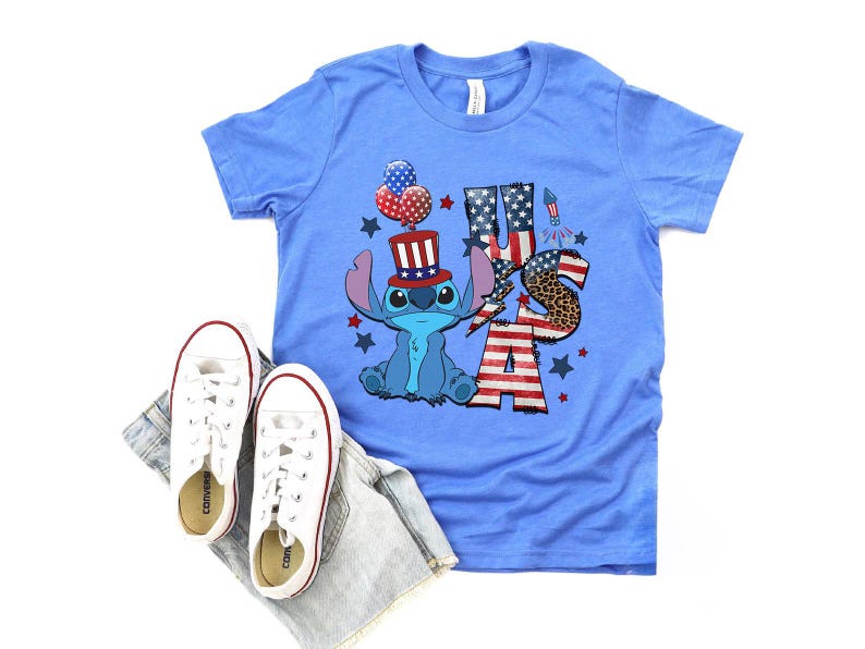 K&ouml;nnte beinhalten: Hellblaues T-Shirt mit einer Cartoonfigur, die einen patriotischen Hut tr&auml;gt und das Wort "USA" in Rot, Wei&szlig; und Blau zeigt. Wei&szlig;e Turnschuhe und Jeansshorts liegen neben dem Shirt.