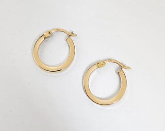 May include: Two gold-colored hoop earrings. The earrings are round with a squared-off profile. They are displayed on a white, oval-shaped tray. The background is a soft pink fabric.