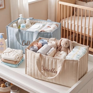 May include: Two rectangular quilted fabric storage caddies, one light blue and one cream, each featuring a name embroidered in brown script. The cream caddy contains rolled baby clothes, a plush rabbit toy, and a stack of nappies. The blue caddy holds baby care bottles and folded cloths.