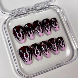 Può includere: Un set di dieci unghie finte a forma di mandorla. Ogni unghia presenta una base rosso scuro con disegni di fiamme bianche e una punta rosa. Piccoli strass trasparenti sono posizionati su ogni unghia. Le unghie sono esposte in una custodia di plastica trasparente.
