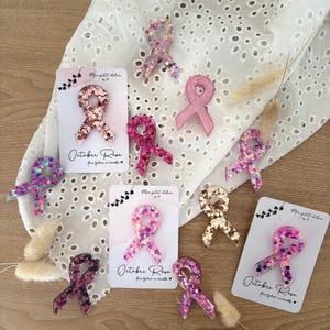 Peut inclure: Plusieurs broches en forme de ruban rose, avec des paillettes et des surfaces texturées. Certaines sont présentées sur des cartes avec le texte "Octobre Rose". Les broches varient en nuances de rose et de violet, avec des paillettes dorées.