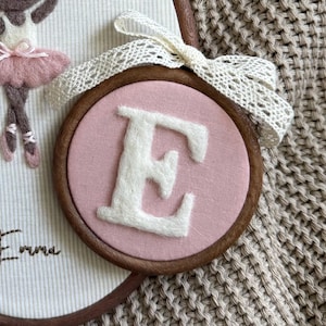 May include: A circular wooden embroidery hoop containing a dusty pink fabric background with a large, white, felted letter E centered. A decorative cream-colored lace ribbon is tied in a bow at the top of the hoop frame, adding a textured detail.