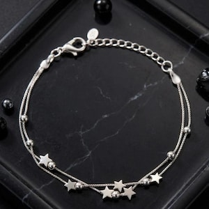 May include: A silver-colored bracelet with a delicate chain design. The bracelet features small star-shaped charms and spherical beads. The bracelet is displayed on a black surface, showcasing its intricate details and design.