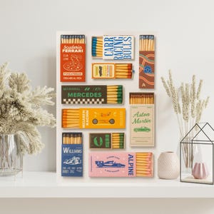 May include: A selection of matchboxes with retro designs, showcasing racing car themes. The boxes are in diverse colours, including red, blue, green, and pink, with text like "Ferrari", "Mercedes", and "Alpine". The matches are visible.