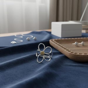 Puede incluir: Un anillo de plata con forma de flor y centro dorado es el punto focal de esta imagen, sobre una tela azul oscuro. También se ven otras joyas, como un collar y pendientes. Una bandeja de madera y una caja blanca están en el fondo.