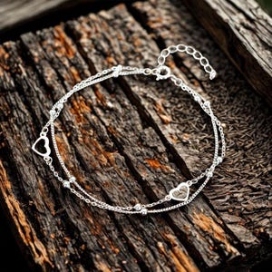 May include: A silver double-strand bracelet with heart and bead accents. The bracelet is made of delicate chains and features two heart-shaped charms and small beads. The bracelet is displayed on a textured wooden surface.