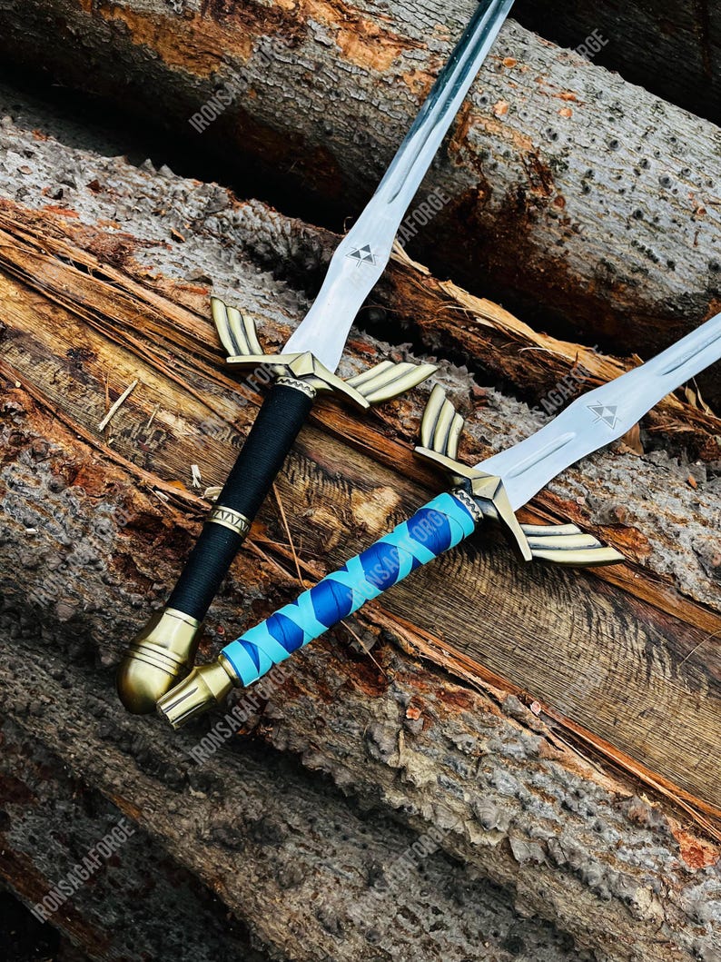 May include: Two decorative swords with silver blades and gold-colored crossguards. One sword has a black handle, and the other has a blue and light blue wrapped handle. The swords are displayed on a wooden surface. The text "IRONSAGAFORGE" is visible.
