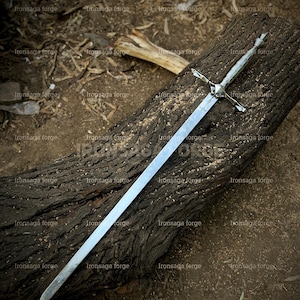 K&ouml;nnte beinhalten: Ein langes, silbernes Schwert mit kreuzf&ouml;rmigem Knauf liegt auf einem verwitterten, dunkelbraunen Baumstamm. Die Klinge ist gerade und poliert und reflektiert das Licht. Der Knauf des Schwertes weist komplizierte Details auf. Der Hintergrund ist nat&uuml;rlich und erdig.