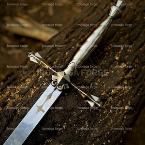 K&ouml;nnte beinhalten: Ein silbernes Schwert mit kreuzf&ouml;rmigem Knauf und langer, reflektierender Klinge. Das Schwert liegt auf einer dunkelbraunen, strukturierten Holzoberfl&auml;che. Die Klinge hat ein ge&auml;tztes Dekor.