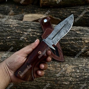 May include: A Damascus steel hunting knife with a dark wooden handle and a leather sheath. The blade has a unique pattern, and the handle is riveted with brass pins. The knife is displayed against a backdrop of wooden logs.