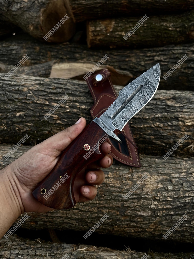 May include: A Damascus steel hunting knife with a dark wooden handle and a leather sheath. The blade has a unique pattern, and the handle is riveted with brass pins. The knife is displayed against a backdrop of wooden logs.