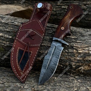 May include: A hunting knife with a patterned blade and a wooden handle, next to a brown leather sheath. The knife has a silver bolster and a decorative pin on the handle. The sheath has a flap closure.