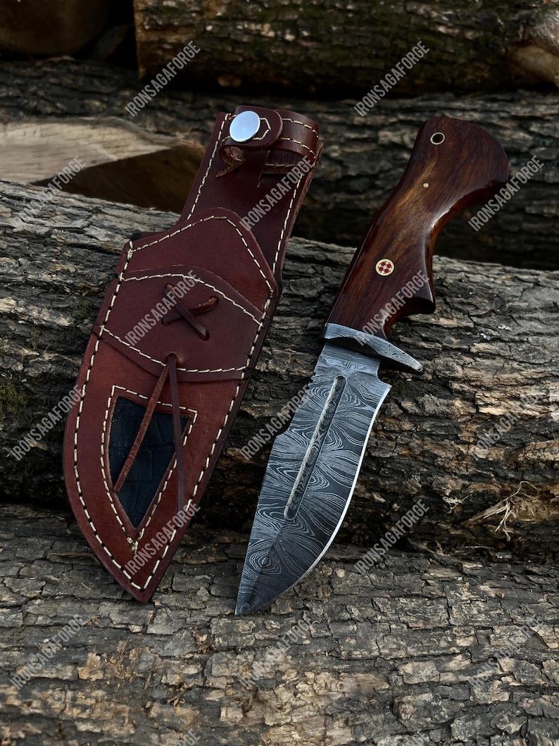 May include: A hunting knife with a patterned blade and a wooden handle, next to a brown leather sheath. The knife has a silver bolster and a decorative pin on the handle. The sheath has a flap closure.