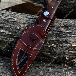 May include: A hunting knife with a dark wooden handle and a brown leather sheath. The knife features a silver bolster and a decorative pin. The sheath is stitched with white thread and has a leather tie. The knife is resting on a wooden surface.