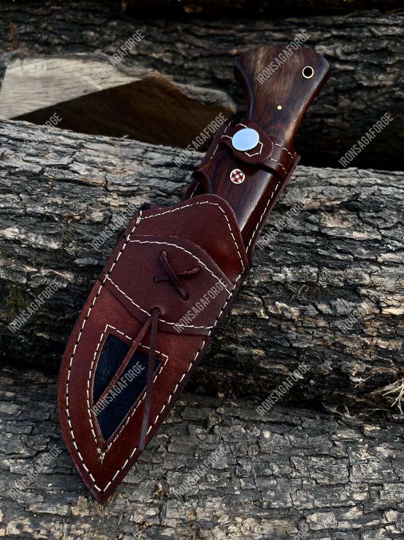 May include: A hunting knife with a dark wooden handle and a brown leather sheath. The knife features a silver bolster and a decorative pin. The sheath is stitched with white thread and has a leather tie. The knife is resting on a wooden surface.