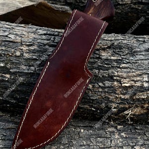 May include: A handcrafted hunting knife with a dark wooden handle and a brown leather sheath. The sheath is stitched with white thread and features a belt loop. The knife is designed for outdoor use and survival.