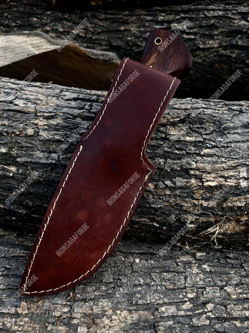 May include: A handcrafted hunting knife with a dark wooden handle and a brown leather sheath. The sheath is stitched with white thread and features a belt loop. The knife is designed for outdoor use and survival.