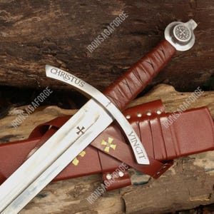 Puede incluir: Una espada medieval con una hoja plateada y un mango envuelto en cuero marrón. La hoja tiene grabadas las palabras "CHRISTUS" y "VINCIT". La espada está en una vaina de cuero marrón con detalles de cruz dorada.