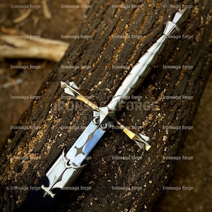 K&ouml;nnte beinhalten: Detailliertes Bild eines mittelalterlichen Schwertes mit silberner Klinge und Griff, mit aufwendigen Designs. Das Schwert liegt auf einer strukturierten, dunkelbraunen Holzoberfl&auml;che. Die Klinge hat einen blauen Farbton, und der Griff hat ein kreuzf&ouml;rmiges Design.