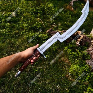 May include: A curved, silver-colored sword with a wooden handle is held against a backdrop of green foliage. The blade features decorative etchings and star-shaped accents. The handle is a dark brown color with silver accents. The text "IRONSAGAFORGE" is visible.