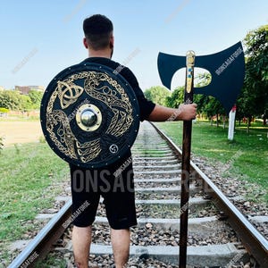 May include: A person stands on train tracks, holding a black shield with gold Celtic knotwork and a large black axe. The axe has a wooden handle and a gold-colored accent. The shield has a central metal boss.