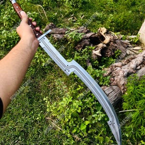 May include: A curved, silver blade sword with a wooden handle. The blade has decorative star-shaped cutouts and a detailed pattern along the spine. The handle is a dark brown color with silver rivets. The sword is held against a backdrop of green grass and foliage.