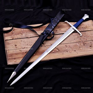 May include: A long, silver sword with a blue handle and crossguard lies on a wooden surface beside a black sheath. The sword has a double-edged blade and a pointed tip. The sheath is made of black leather and has a strap.