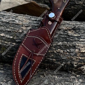 K&ouml;nnte beinhalten: Ein handgefertigtes Jagdmesser mit einem dunklen Holzgriff und einer braunen Lederscheide. Das Messer hat einen silbernen Knauf und einen dekorativen Stift. Die Scheide ist mit wei&szlig;em Faden gen&auml;ht und hat eine schwarze Innenseite. Messer und Scheide liegen auf einer Holzoberfl&auml;che.