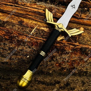 May include: A decorative sword with a silver blade and gold-colored hilt. The hilt features a black wrapped handle and gold accents. The sword is displayed on a wooden surface.