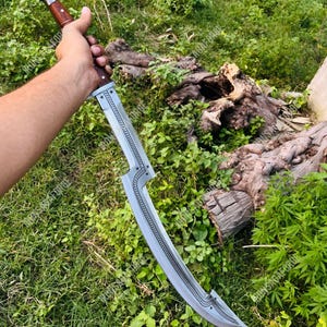 May include: A curved, silver-colored blade with a wooden handle. The blade has a decorative star pattern and a curved design. The handle is a dark brown color. The sword is held against a backdrop of green foliage.