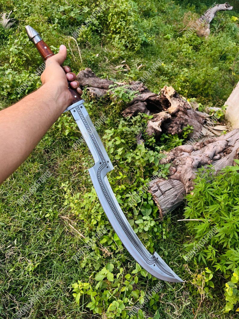 May include: A curved, silver-colored blade with a wooden handle. The blade has a decorative star pattern and a curved design. The handle is a dark brown color. The sword is held against a backdrop of green foliage.