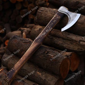 May include: A handcrafted axe with a polished steel blade and a dark wooden handle. The handle features etched runes and the text "Ironsaga Forge". The axe is displayed on a pile of wooden logs, showcasing its rustic design.
