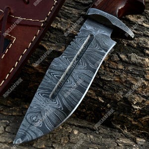 May include: A Damascus steel knife with a wooden handle and a leather sheath. The blade has a unique pattern, and the handle is dark brown. The sheath is a lighter brown with stitching and a leather strap. The text "IRONSAGAFORGE" is visible.