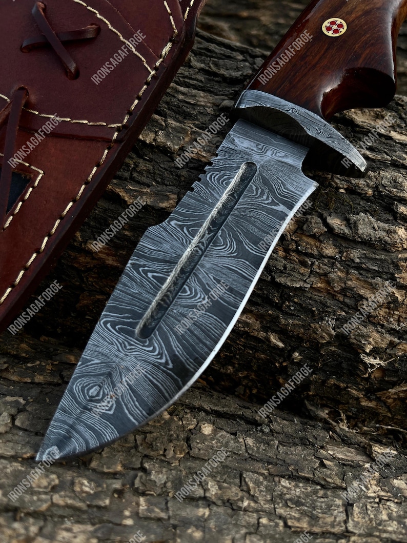 May include: A Damascus steel knife with a wooden handle and a leather sheath. The blade has a unique pattern, and the handle is dark brown. The sheath is a lighter brown with stitching and a leather strap. The text "IRONSAGAFORGE" is visible.