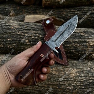 K&ouml;nnte beinhalten: Ein Jagdmesser aus Damaststahl mit einem dunklen Holzgriff und einer Lederscheide. Die Klinge hat ein einzigartiges Muster, und der Griff ist mit Nieten befestigt. Das Messer wird vor einem Hintergrund aus Holzst&auml;mmen gezeigt.