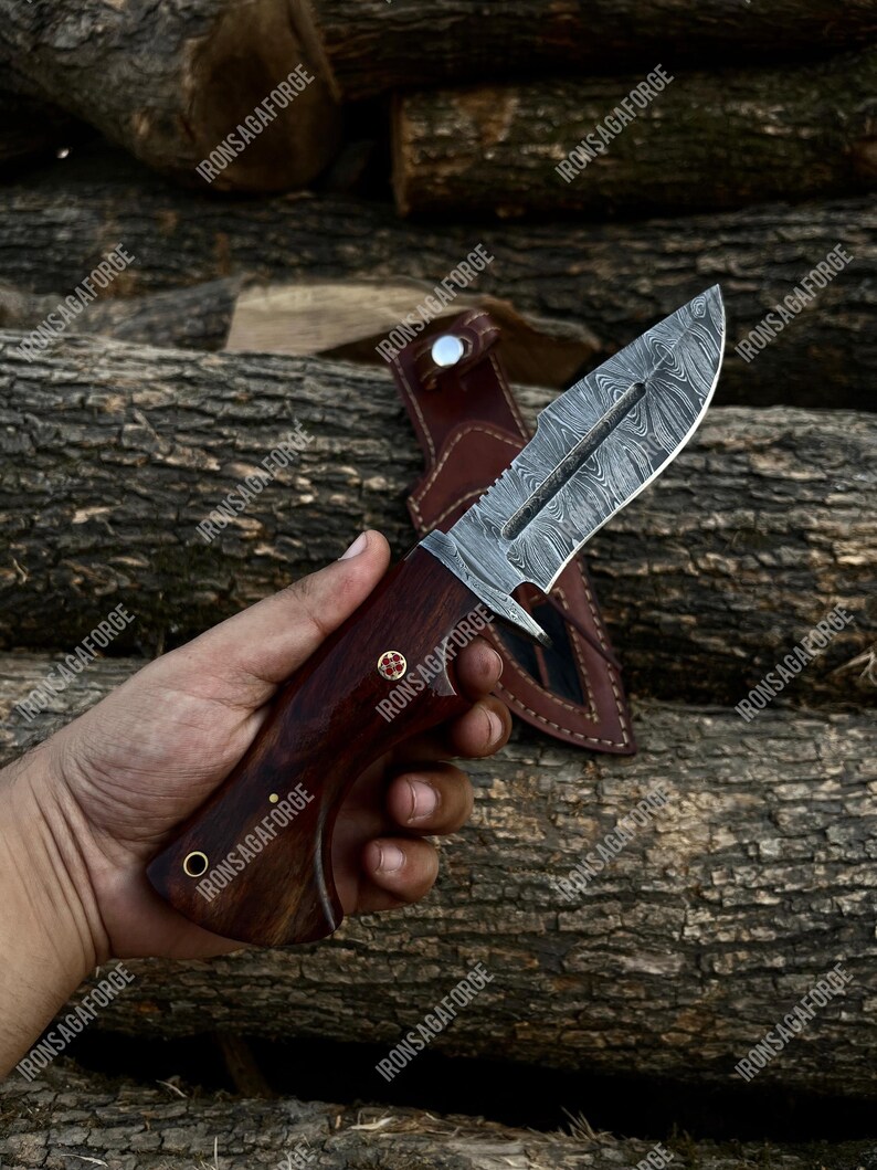 K&ouml;nnte beinhalten: Ein Jagdmesser aus Damaststahl mit einem dunklen Holzgriff und einer Lederscheide. Die Klinge hat ein einzigartiges Muster, und der Griff ist mit Nieten befestigt. Das Messer wird vor einem Hintergrund aus Holzst&auml;mmen gezeigt.