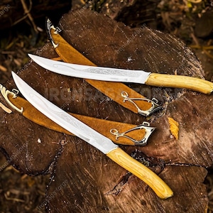 Könnte beinhalten: Zwei Messer mit silbernen Klingen, Holzgriffen und dekorativen Scheiden. Die Klingen haben geätzte Designs, und die Griffe sind hellholzfarben. Die Scheiden sind aus braunem Leder mit goldfarbenen Metallakzenten.