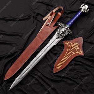 May include: A detailed image of a fantasy sword with a silver blade and a blue handle. The sword features a lion's head design on the hilt and rests beside a wooden shield with gold accents. A brown leather sheath is also present.