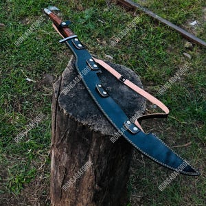 May include: A large, curved blade sword with a brown handle and a black leather sheath. The sheath has white stitching and is secured with straps. The sword rests on a tree stump against a backdrop of green grass.