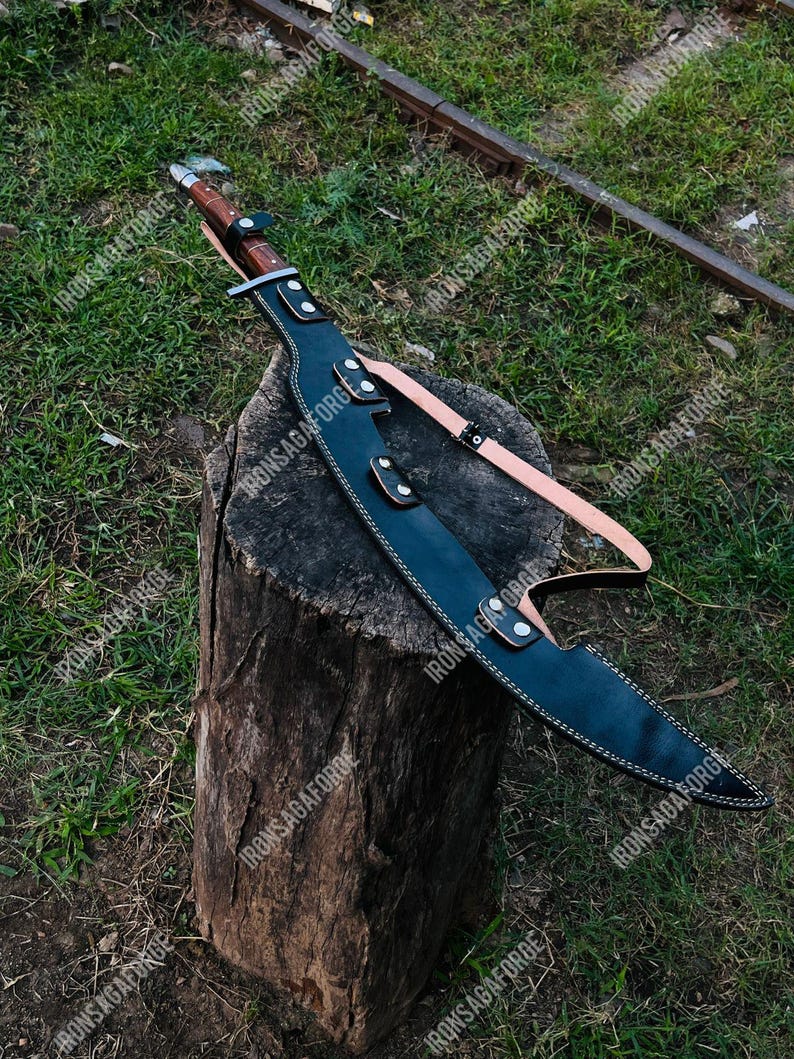May include: A large, curved blade sword with a brown handle and a black leather sheath. The sheath has white stitching and is secured with straps. The sword rests on a tree stump against a backdrop of green grass.