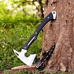 May include: A black and silver axe with a curved handle and a toothed blade, resting against a tree trunk. The axe has a protective sheath. The background features green foliage and a wooden stump.