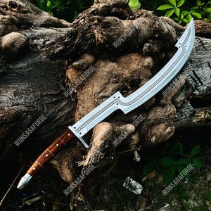 May include: A curved, silver-colored sword with a wooden handle. The blade has a decorative etched design and is set against a natural wood background. The handle is a dark brown color with silver accents. The sword is a fantasy or historical weapon.