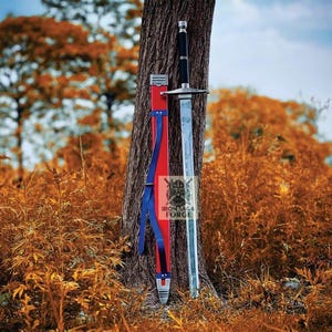 May include: A long sword with a black handle and silver blade, leaning against a tree. The sword is next to a red and blue sheath. The background features a blurred autumn landscape with orange and brown foliage.
