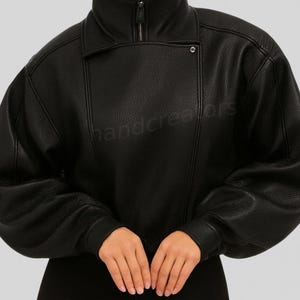 Op de afbeelding: Zwarte leren jas met een hoge kraag en ritssluiting. De jas heeft een getextureerde uitstraling en een knoopdetail aan de voorkant. De mouwen zijn lang en licht gezwollen. De tekst "handcreators" is zichtbaar.